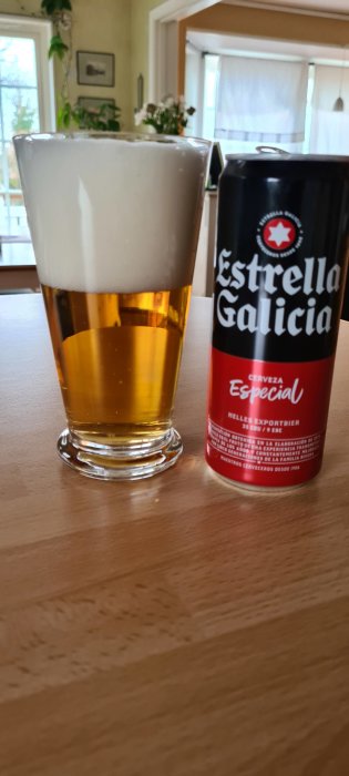 Ett glas med öl och en burk av Estrella Galicia på ett bord inomhus.