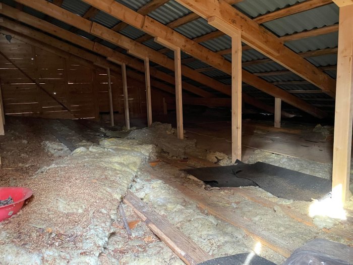Oisolerat vindsutrymme med exponerad isolering, trästolpar och sprickor i svart takmaterial.