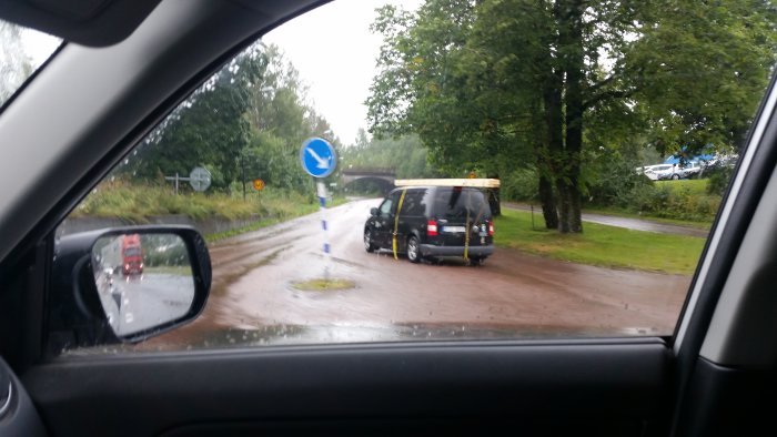 Översvämmad vägkorsning med bil och trafikskyltar sedd från bilfönster under regnig dag.