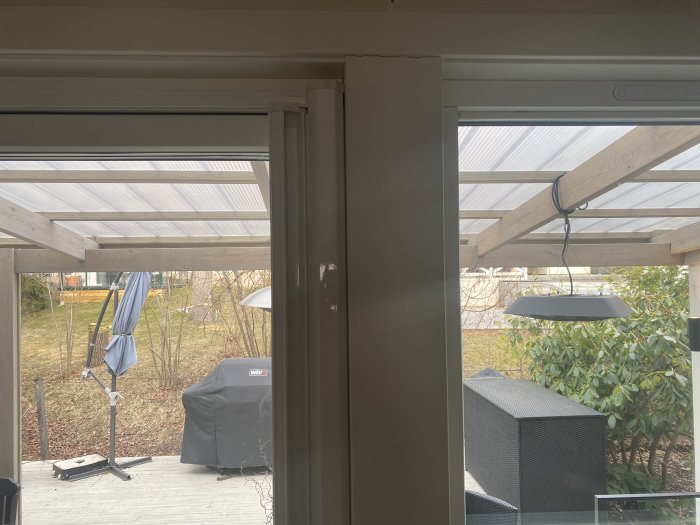 Utsikt från ett fönster mot en halvbyggd pergola med transparent tak och trädgårdsmöbler.