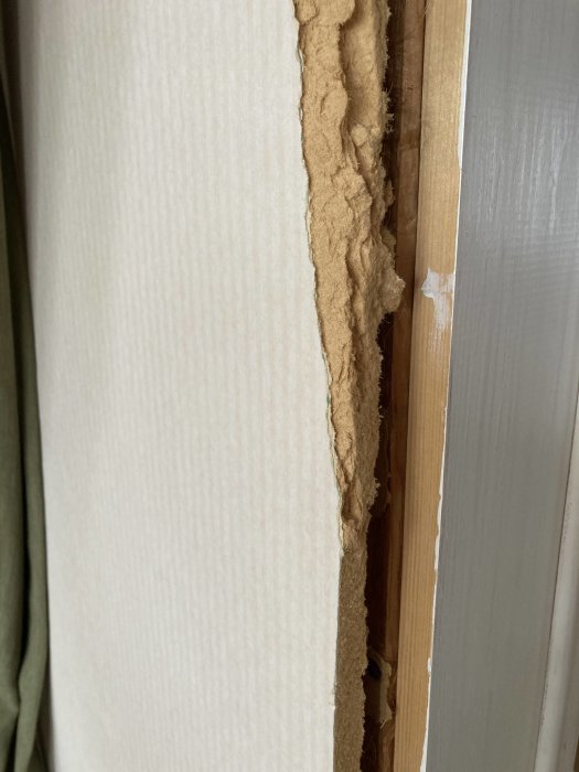 Skadad tapet avslöjar underliggande brunt material, möjligen isolering, nära trälist.