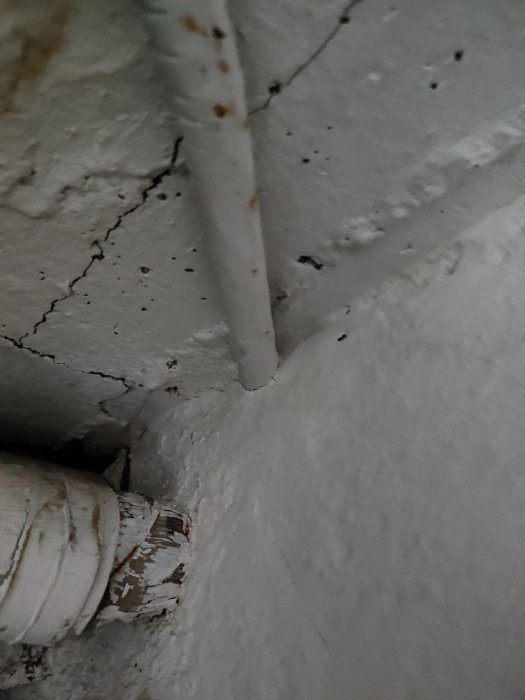 Äldre rörledning som är fast i en sprucken och flagnande vitmålad vägg, tecken på slitage från mitten av 1900-talet.
