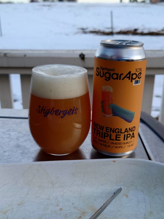 Ölglas med skum fyllt med New England Triple IPA bredvid en tom ölburk på ett bord utomhus med snö i bakgrunden.