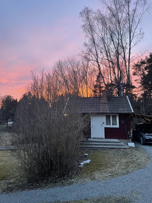 Ett litet rött hus vid solnedgång med träd och en bil i förgrunden.