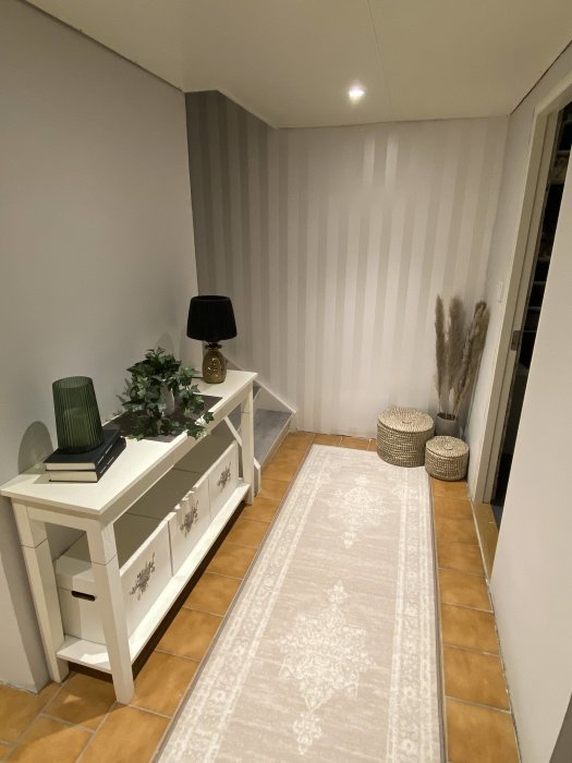En oinredd källarhall med en vit konsolbord, lampa, matta och terrakottafärgad klinker.