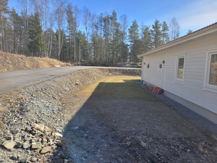 Vy över en slänt längs med ett hus, med bar jord och några spridda stenar, redo för landskapsarbete.