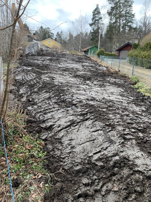 En sluttande tomt täckt med lera och hjulspår, omringad av träd och hus i bakgrunden.