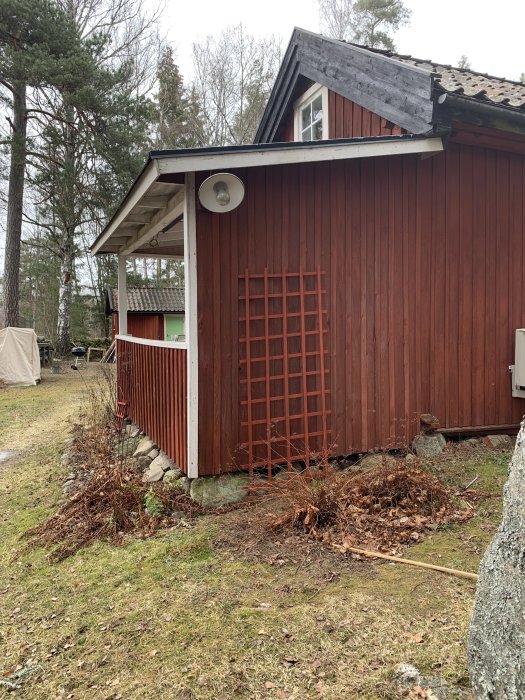 Tom spaljé mot en röd husvägg i en trädgård med träd som ger skugga.