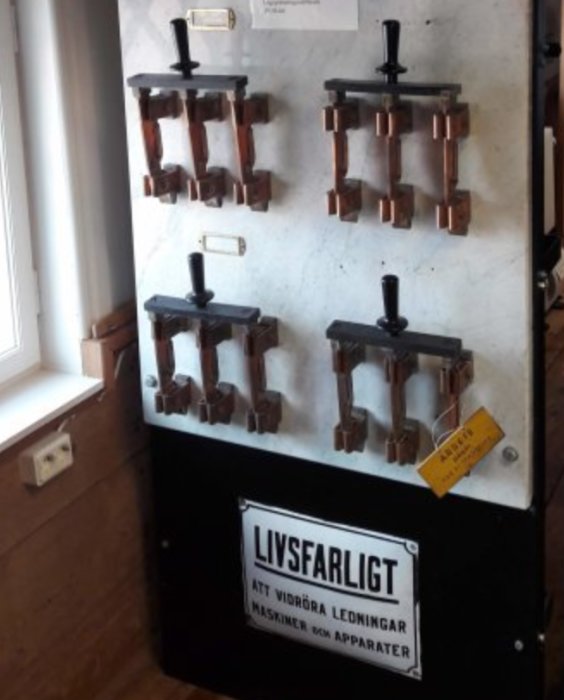 Äldre elektrisk kopplingstavla med skylten "Livsfarligt att vidröra ledningar" på en svart bakgrund.