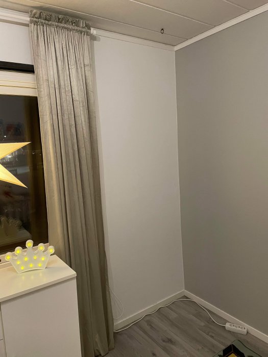 Ny målad grå vägg i ett barnrum med ljusgrå gardin och en ljuskrona formad som en krona.