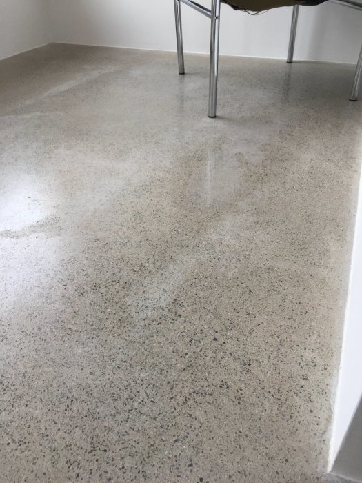 Slipat betonggolv med synliga fläckar och ojämnheter, del av möbelben i övre högra hörnet.