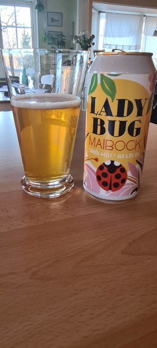 Ölglas med gul vätska bredvid burk märkt "LADY BUG MAIBOCK", gult och rosa, på träbord.