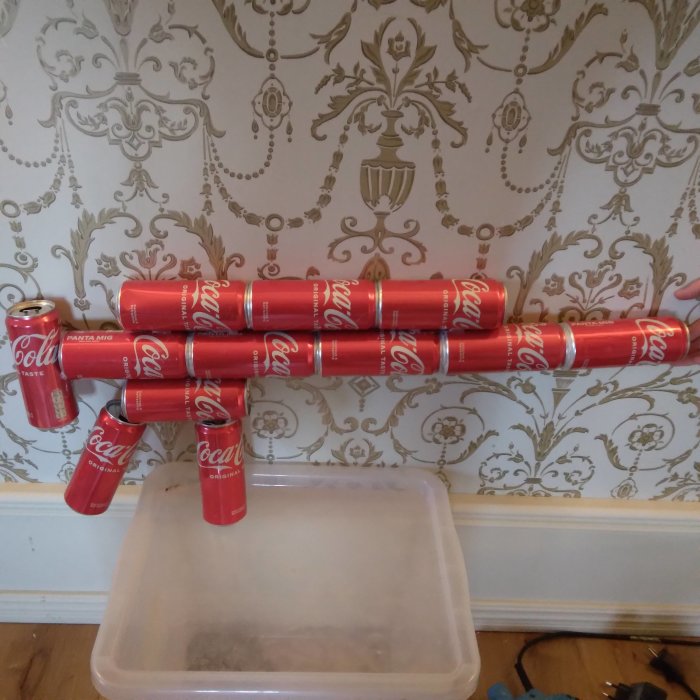 Hemmagjord hylla av Coca-Cola-burkar framför en vägg med tapet, en plastlåda under som skydd.