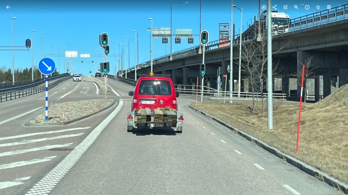 Röd skåpbil med träplankor säkrade av spännband kör på väg vid avfart, klarblå himmel.