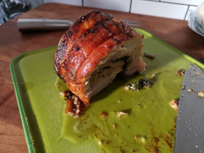 Delvis skivad porchetta med krispig yta på ett grönt skärbräde bredvid en kökskniv.