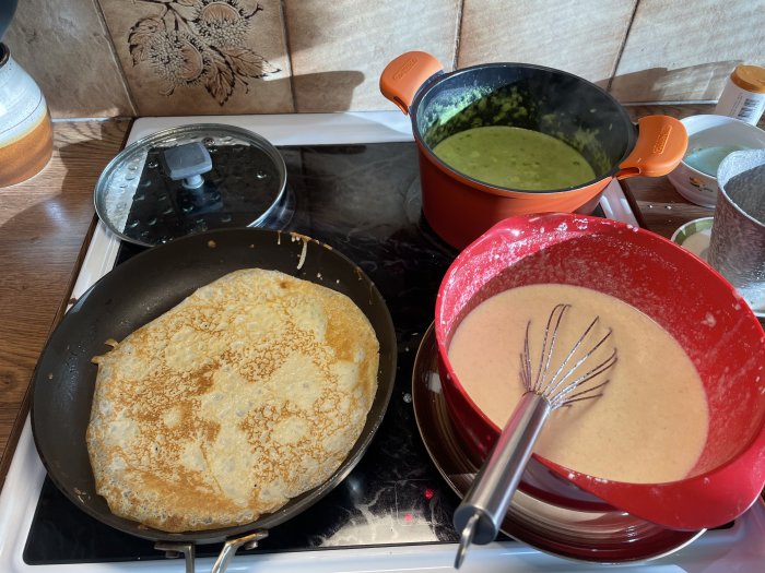 Ärtsoppa i orange kastrull och pannkakssmet i röd skål med visp bredvid en stekpanna med pannkaka.