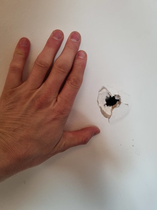 Hand jämför storleken på ett hål i en vit gipsvägg som behöver repareras.