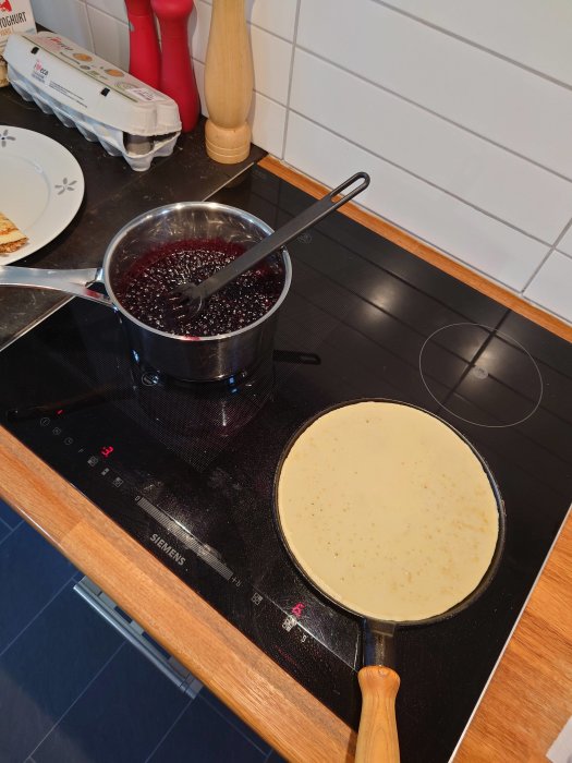 En pannkaka steks i stekpanna på spis bredvid kruka med blåbärssylt.