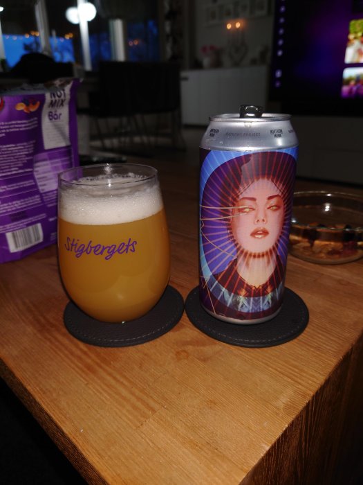 Ölglas med skum på träbord och en burk öl med konstnärlig etikett.