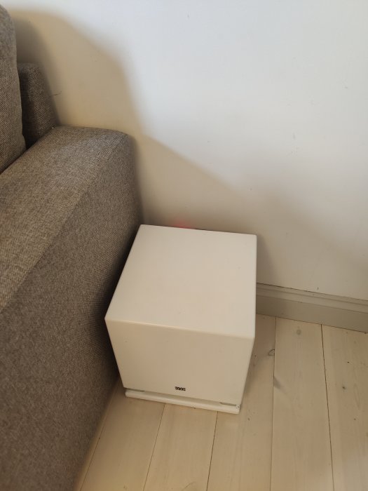 Vit centerhögtalare placerad på ett trägolv bredvid en soffas gråa tygkant.