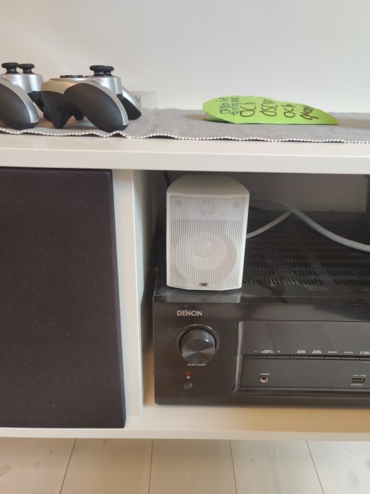 Centerhögtalare på hylla ovanför en AV-mottagare från Denon, flankerad av spelkontroller.