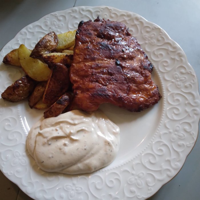 Grillad fläsknoisette med klyftpotatis och örtagårdssås på en vit tallrik.