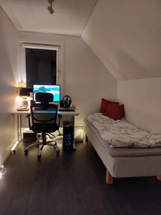 Kompakt hemmakontor med dator, skrivbordsstol och dagbädd under snedtak, kvällsbelysning.