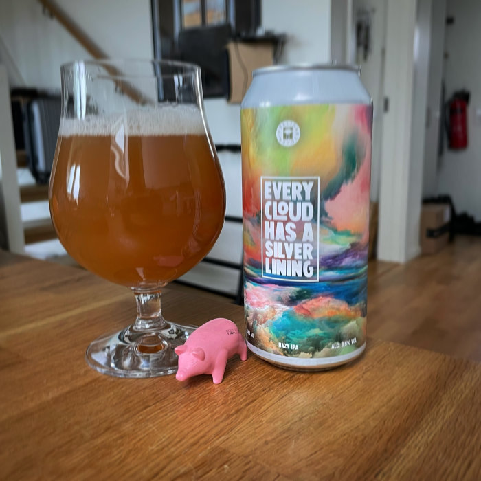 Ölglas med bärnstensfärgad dryck och färgglad ölburk med texten "Every Cloud Has a Silver Lining", rosafärgad plastgris framför.