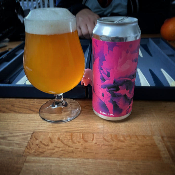 Ett glas fyllt med orange öl bredvid en burk Double IPA med rosa abstrakt design på ett träbord.