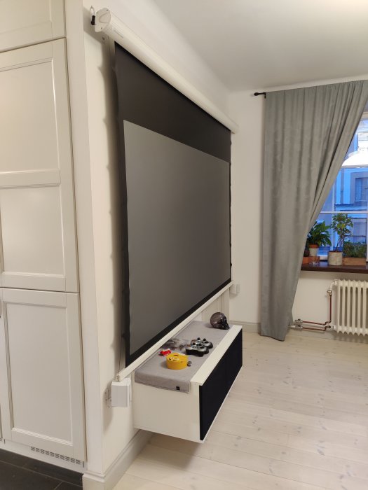 Projektorduk delvis nere med synliga ytterkanter, satt över vit möbel med spelkontroller, i ett vardagsrum.