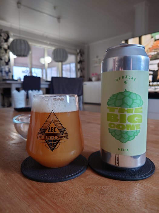 Ölglas med logotyp framför en burk öl på ett bord i ett kök.