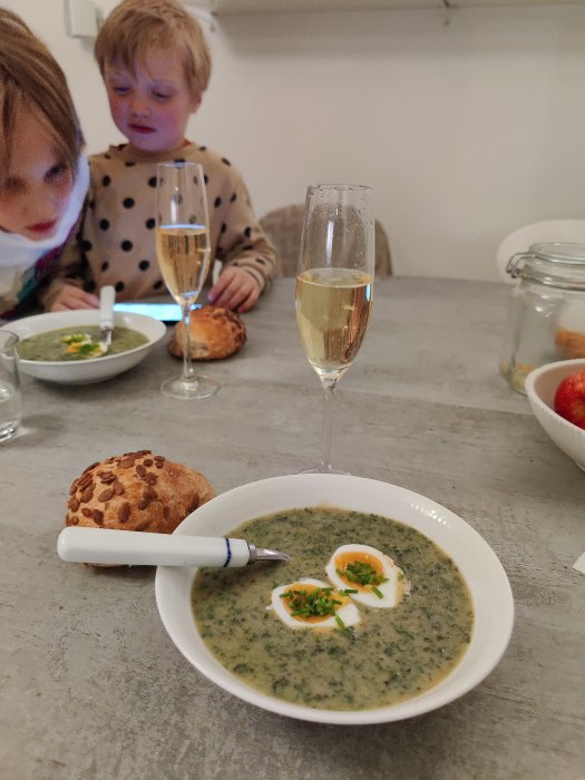 Ett glas Riesling sekt bredvid en skål med grönsakssoppa och ett runt bröd, med två barn i bakgrunden.