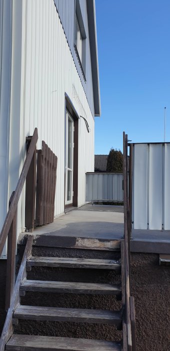 Balkong ovanpå garage med halvtäckt plåtfasad och trätrappa mot en vitputsad husvägg.