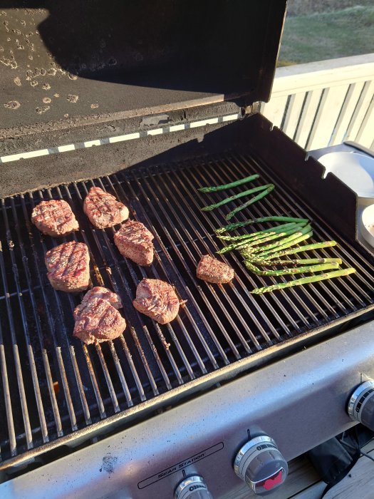 Stekta biffar och gröna sparris på gasgrillens varma galler i solljus.