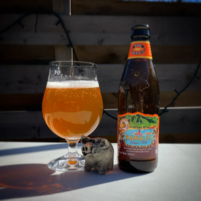 Ölglas fyllt med öl bredvid en flaska av Hanalei Island IPA och en liten lerfigur på ett bord i solljus.