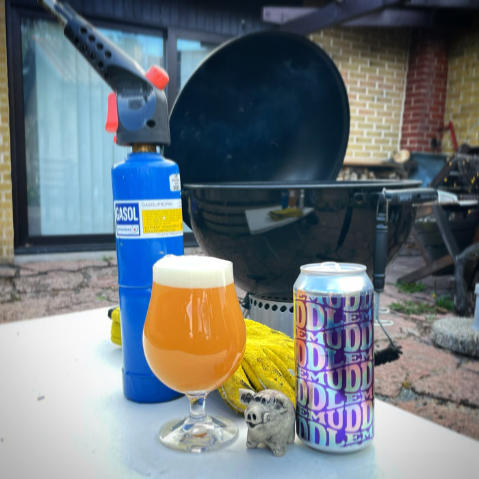 En ölglas fylld med öl framför en gasolbrännare, grill och en ölburk med märklig design.