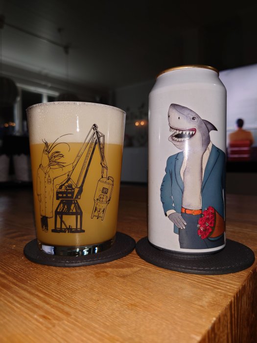 Ölglas med Göteborgsmotiv och ölburk med haj i kostym och rosor, på träbord.