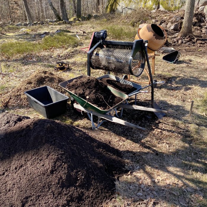 Byggplats med sikttumlare i arbete och siktad jord, skottkärror, hög av jord och skogsmark i bakgrunden.