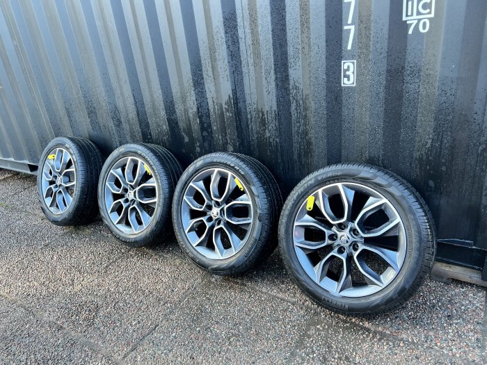 Fyra bilfälgar med däck lutade mot en container på en grusunderlag.