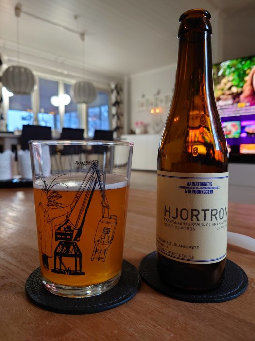 Ett glas med gyllene dryck och en flaska märkt Hjortron från Mariatorgets Mikrobryggeri på ett bord.