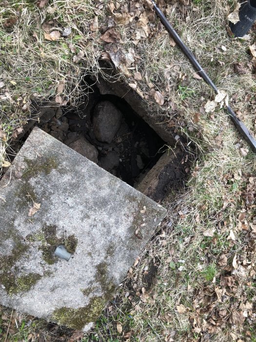 Betonblock med rörhandtag och sikt in i hålet i marken med stenar och gammal ved.