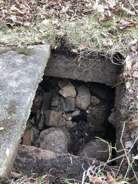 Gamla stenar och ved i en 1 meter djup grop under ett betongblock med grusballast och rörhandtag.