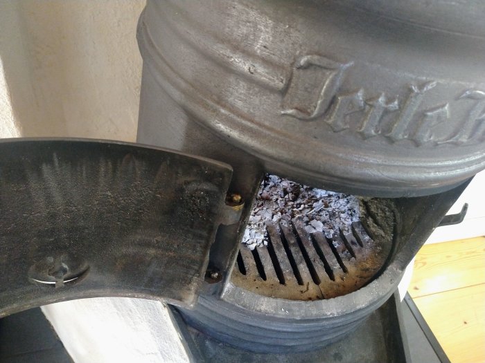 Öppen kaminlucka med synliga gångjärnstappar och aska i brännkammaren, tips om smörjning med kopparpasta.