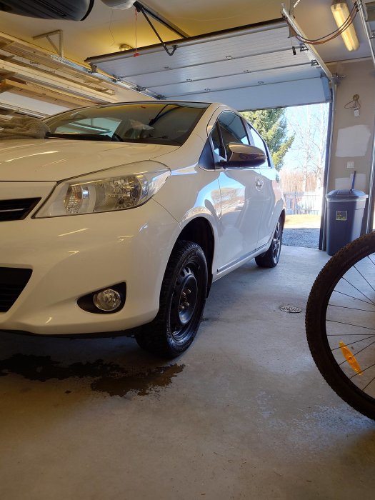 Vit bil parkerad i ett garage med en cykel framför och en del av garaget synligt.