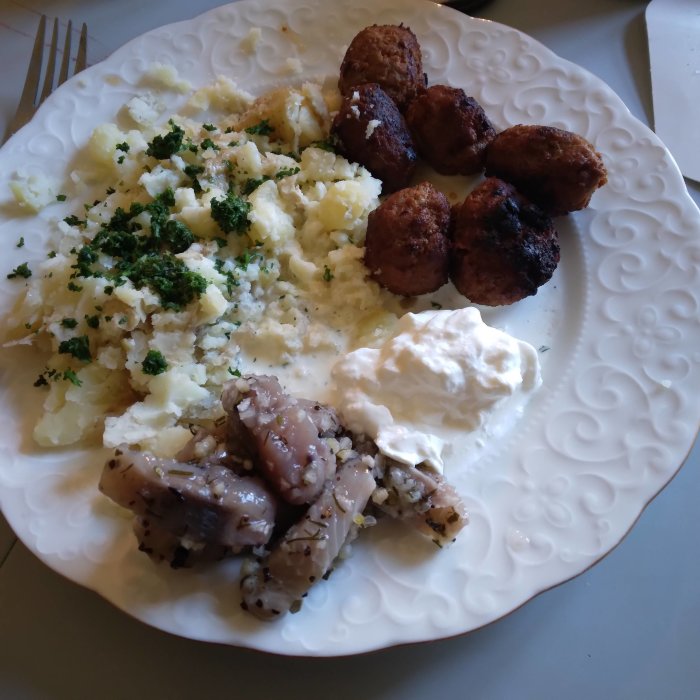 Svensk middagstallrik med sill, potatismos, köttbullar och gräddfil, dekorerad med hackad persilja.