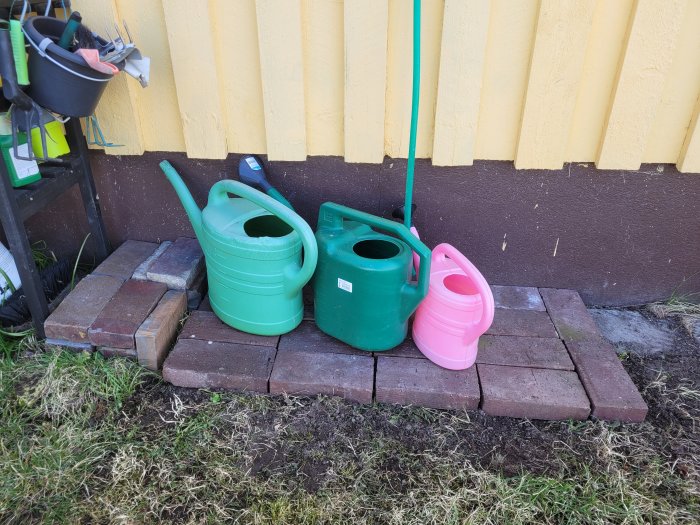 Två gröna och en rosa vattenkanna uppställda på en stadig bas av marksten vid en gul husvägg.