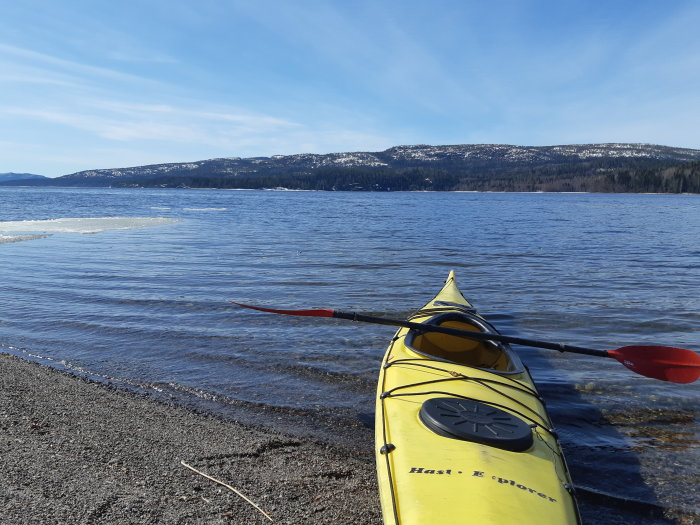 Gul kajak med paddel vid strandkanten, snötäckta berg och isflak i bakgrunden.