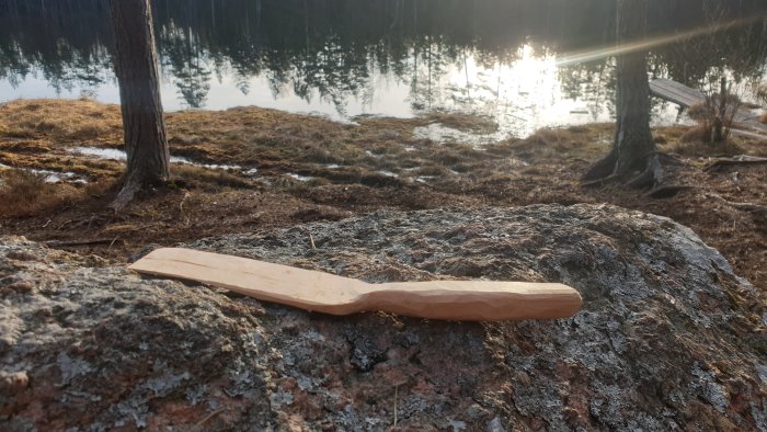 Handtäljd trästekspade på en sten framför en sjö och skog i vårljuset.