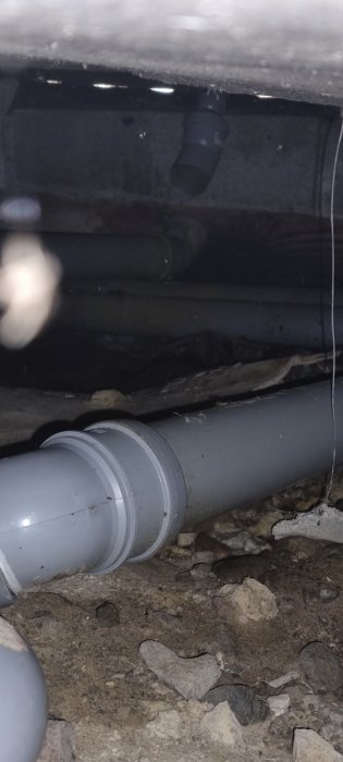 Rörledningar under ett hus med grus och stenar synliga under golvbjälklaget, dålig belysning.
