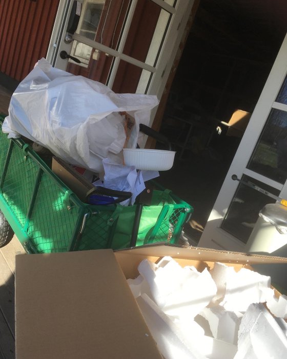 Grön shoppingvagn full med vita soppåsar, frigolitbitar och kartonger vid en öppen dörr med insyn till en loge.